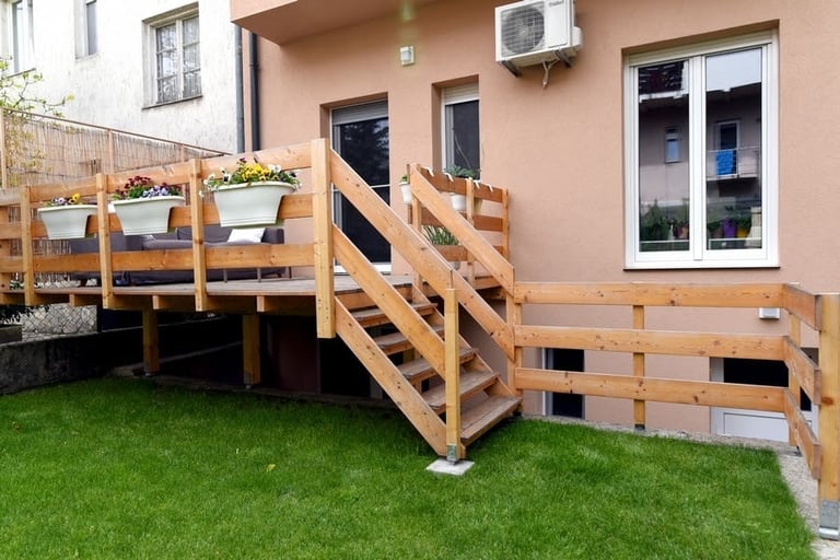 Wooden deck with stairs and railings built on green lawn in front of a peach-colored house with potted plants